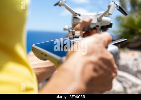 Close-up of hands operating a remote controller and smartphone while preparing a quadcopter drone for flight outdoors, with blue sea and rocky coast b Stock Photo