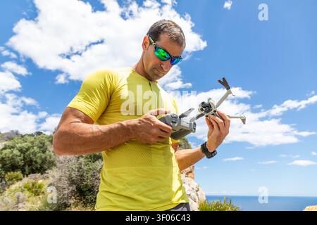 Athletic man in yellow sport shirt and sunglasses adjusts a drone controller while holding a quadcopter outdoors on a sunny day near cliffs and ocean. Stock Photo