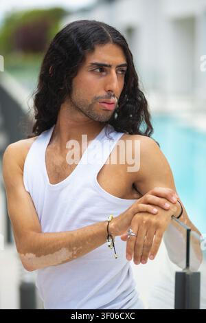 Middle Eastern man with vitiligo stands by a swimming pool, wearing a white tank top and looking away. Outdoor lifestyle portrait highlighting skin di Stock Photo