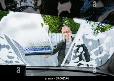 View from inside a vehicle as a man cleans the windshield with a brush and thick soap foam at a self service car wash, outdoor setting, routine vehicl Stock Photo