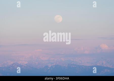 Soft pastel moon hangs above the Alps at dawn, with layered snowy ridges and misty clouds fading into a tranquil horizon, creating a calm, dreamy moun Stock Photo