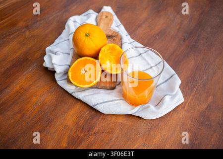 A refreshing glass of freshly squeezed orange juice sits next to whole and halved oranges on a rustic wooden table, accompanied by a striped cloth, ca Stock Photo