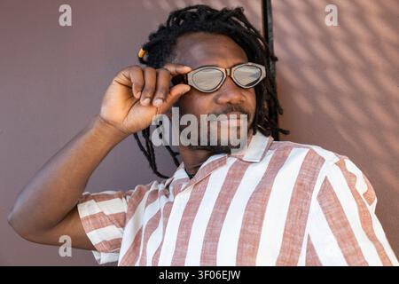 Portrait of a Cuban man with dreadlocks adjusting stylish sunglasses, wearing a striped button-up shirt in warm sunlight against a textured wall, rela Stock Photo