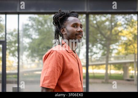 Side profile portrait of a Cuban man with dreadlocks in an orange shirt, standing outdoors near a modern glass building, looking ahead in soft natural Stock Photo