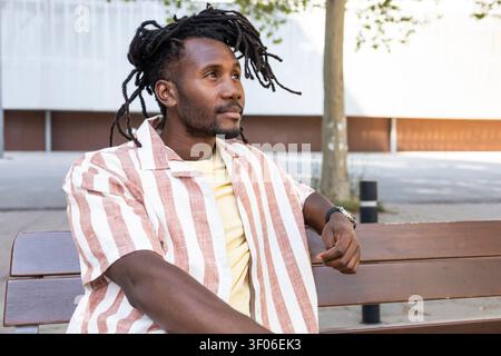 Portrait of a Cuban man with dreadlocks relaxing on a wooden bench in an urban park, looking away thoughtfully in soft daylight, casual striped shirt Stock Photo
