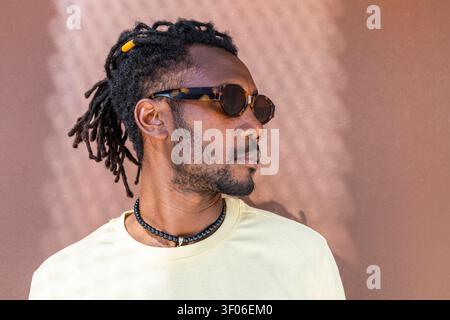 Side profile portrait of a Cuban man wearing sunglasses, dreadlocks, and a beaded necklace, standing against a warm textured wall with natural light. Stock Photo