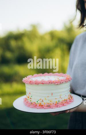 Close-up of a person holding a white birthday cake with pink frosting borders and colorful sprinkles, outdoors in natural light with soft bokeh backgr Stock Photo