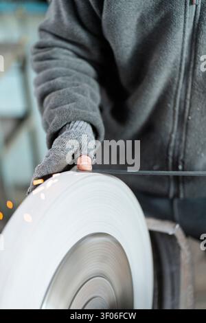 Close-up of a worker wearing gloves using a large abrasive grinding wheel to shape metal, with bright sparks flying in an industrial workshop setting. Stock Photo
