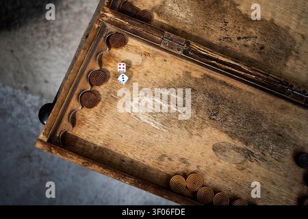 Top view of a worn vintage backgammon board with carved checkers and two dice on rustic wood, set on a dark table, evoking classic strategy gameplay a Stock Photo