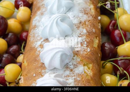 Homemade loaf cake topped with white meringues and powdered sugar, surrounded by red and yellow cherries for a rustic summer dessert still life. Stock Photo