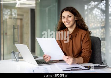 Young businesswoman with curly hair smiling and looking at the camera while holding documents and sitting at her desk with a laptop in a modern office, representing productivity and success Stock Photo