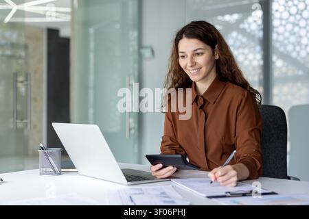 Young businesswoman smiling and working on financial documents at desk in a modern office, using a laptop, calculator, and clipboard for budgeting and data analysis Stock Photo