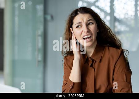 Young woman feeling intense ear pain, holding her painful ear with a grimace, suffering from health problems or hearing issues indoors Stock Photo