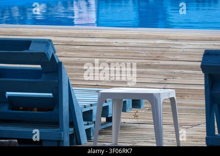 Wooden poolside deck with blue pallet furniture and white side table beside a clear swimming pool, creating a minimalist summer leisure background wit Stock Photo