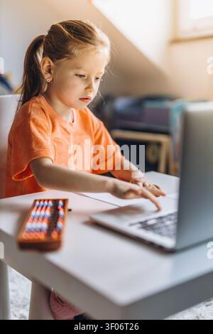 Child practices mental math on an abacus while studying at home with a laptop and learning materials. Mental arithmetic builds concentration, logical Stock Photo