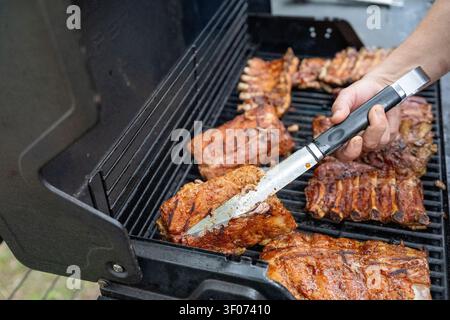 A close-up shot of a person using metal tongs to arrange or flip succulent, glazed pork ribs on a barbecue grill. The meat shows delicious char marks Stock Photo