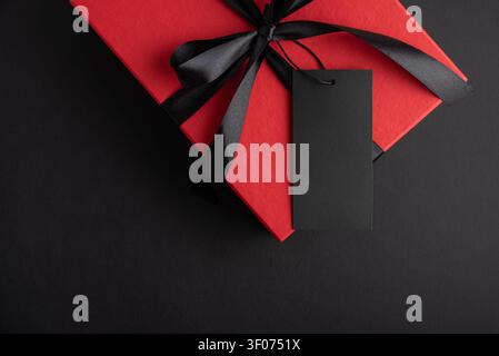 Red textured gift box tied with a black satin ribbon and bow, featuring a blank black tag, resting on a dark background, representing concepts of gift Stock Photo