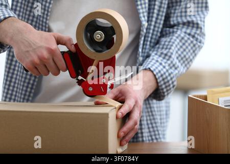 Man using dispenser with adhesive tape to seal cardboard box before sending at table in office, closeup Stock Photo