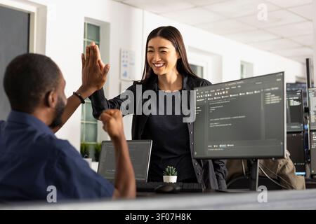 Diverse colleagues celebrate successful project in office, highlighting collaboration, achievement, and positive teamwork. Male and female developers give high fives in startup workspace. Stock Photo