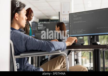 Young adult developer works in wheelchair at modern tech workspace, coding software solutions. Scene highlights disability inclusion, professional IT development and accessible digital innovation. Stock Photo