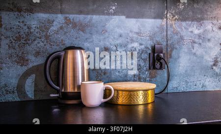 Electric kettle, ceramic mug, and golden storage box with bamboo lid on a stylish dark kitchen counter against a textured blue concrete backsplash. Stock Photo