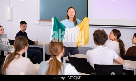 High school teacher tells students about Ireland and holds a Ireland flag in hands. Stock Photo