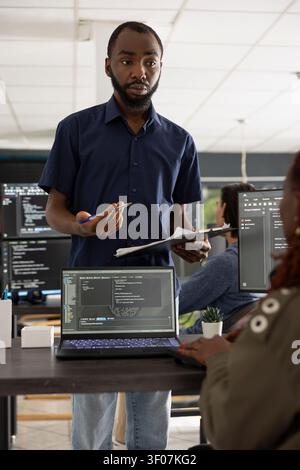 Male manager reviews website progress as coworker types updated software code on desktop pc. The discussion takes place at computer workstation, highlighting collaboration in tech startup office. Stock Photo