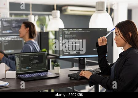 Female software developer taps pen on forehead, studying coding scripts on screen with thoughtful expression. Diligent asian woman works on computer, brainstorming ideas for app development. Stock Photo