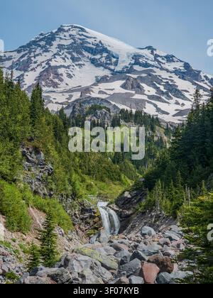 A majestic snow-capped mountain peak rises above a lush green valley with a cascading waterfall and rocky stream bed in Washington state. Stock Photo