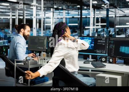 African American woman stretches at a workstation in a server hub to relieve back fatigue after long technical work. Stock Photo