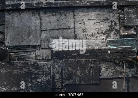 A close-up abstract texture of a patchwork of old weathered wood planks and plywood sections, some painted black, showing peeling finish, England. Stock Photo