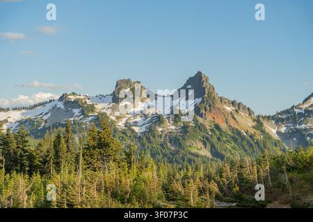 Rugged mountain peaks with lingering snow patches rise above a dense evergreen forest under a clear blue sky in Washington state. Stock Photo