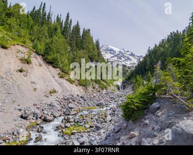 A rocky stream flows through a forested valley with a waterfall and a snow-capped mountain peak in the background. Stock Photo