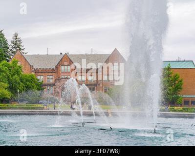 Water jets spray high into the air from a large pond in the foreground with a red brick building and green trees in the background. Stock Photo