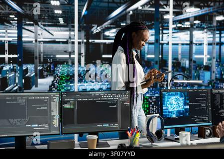 Coder coding in server facility, updating ML automation scripts. African American woman in server facility programming AI algorithms, debugging equipment racks. Stock Photo