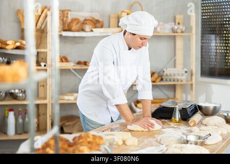 Guy baker in uniform prepares raw dough, kneads it with hands Stock Photo