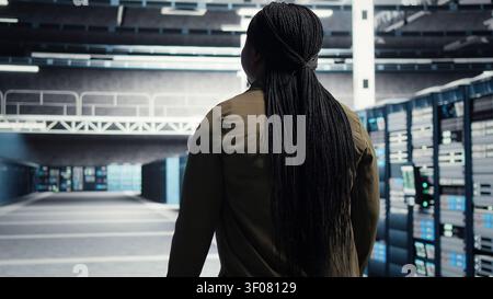 Woman walking through server facility rows of servers, checking equipment for system stability. IT professional supervising server environment, checking infrastructure mainframes. Stock Photo