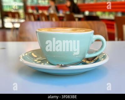 A light blue ceramic cup of latte art sits on a matching saucer with a spoon, placed on a white table in a café. The background shows blurred patrons Stock Photo