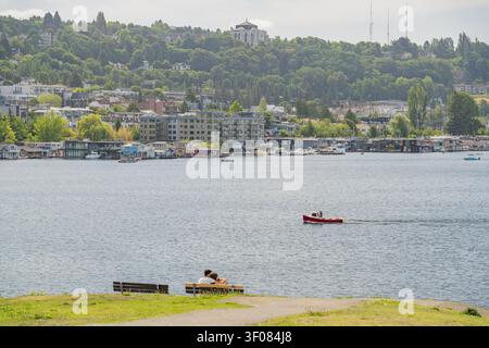 Washington State, JUN 21, 2026 - Two people sitting on a park bench overlooking a body of water with a red boat and city skyline in the background. Stock Photo