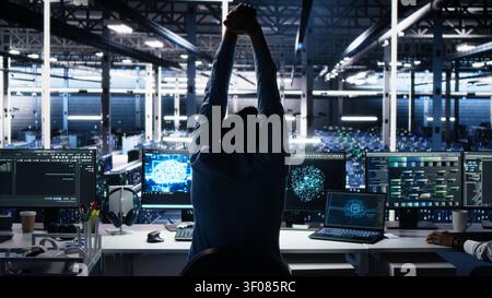 Server room IT technician stretching a sore back while working with AI at desk, while fatigued IT specialist in server facility relaxing strained muscles, feeling backache while using AI tools. Stock Photo
