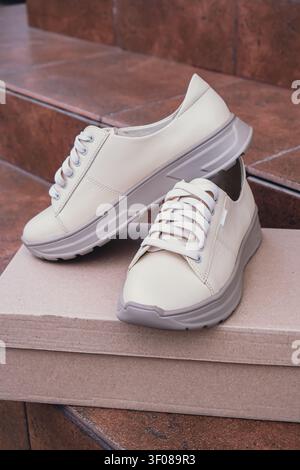 Minimalist eco-friendly white sneakers placed on recycled cardboard box, clean design for sustainable fashion concepts. New unbranded beige sneakers footwear mockups and modern product displays. For branding, e-commerce and packaging design Stock Photo