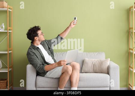 Young man adjusting air conditioner with remote while holding laptop Stock Photo