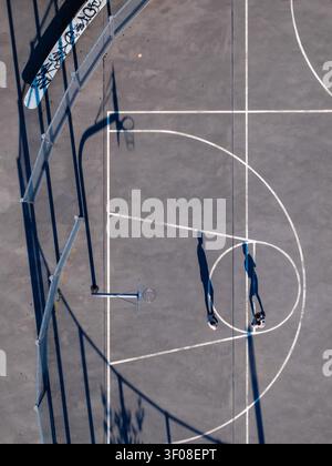 Aerial view of a basketball court with two people casting long shadows on the pavement, surrounded by a fenced area in Mannheim, Baden-Wurttemberg, Ge Stock Photo