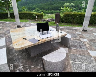 Laptop rests on stone table outdoors. Surrounded by green hills and trees. Peaceful workspace with natural stone furniture. Sunlight enhances tranquil Stock Photo