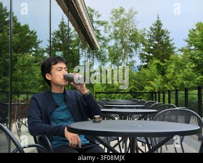 Man sips drink at outdoor cafe table. Green trees frame glassy background. Distant hills add serene depth. Relaxed posture conveys calm leisure. Ideal Stock Photo