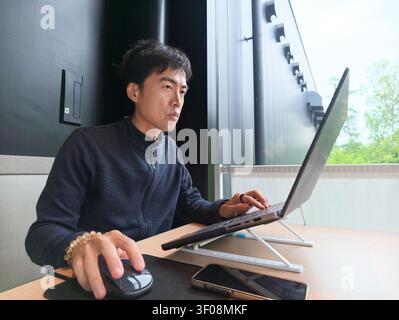 Young man focuses on laptop work. He wears dark sweater and uses mouse. Natural light streams through window behind. Desk holds phone, mouse pad, and Stock Photo