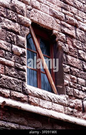 Old wooden window set in massive rustic stone wall of brown blocks, reinforced with diagonal support beam. Architectural detail of historical medieval Stock Photo