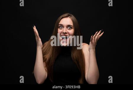 Young woman shouting and expressing excitement, feeling surprised and happy with hands raised on black background Stock Photo