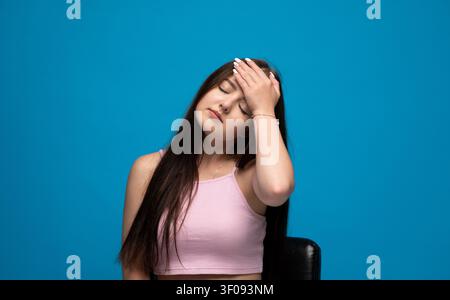 Young woman experiencing severe headache, feeling pain and stress, eyes closed, sitting on chair against blue background Stock Photo