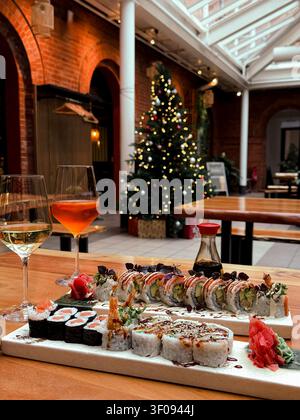 Sushi platter with assorted rolls, soy sauce, and drinks on wooden table, decorated Christmas tree in the background Stock Photo